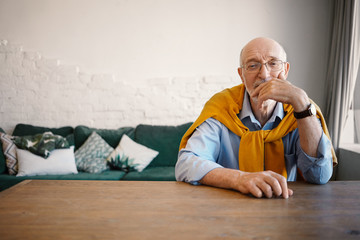 Wall Mural - Indoor shot of handsome elderly mature man with wise eyes sitting at wooden desk with sofa in background, looking at camera with pensive expression, touching face. People, lifestyle and age