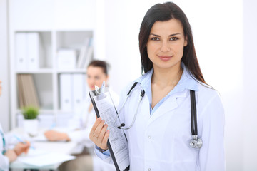 Brunette female doctor on the background of colleagues talking to each other in hospital. Medicine and health care concept