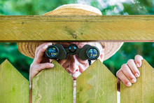 A Curious Neighbor Stands Behind A Fence And Watches With Binoculars
