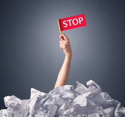 Wall Mural - Female hand emerging from crumpled paper pile holding a red flag with stop written on it 