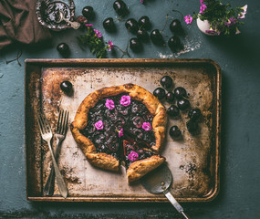 Homemade cherry pie or galette on aged baking tray and rustic kitchen table background with jam and cutlery, top view. Summer berries still life concept