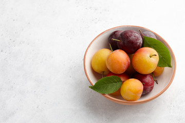 Wall Mural - Fresh colorful plums with leaves in fruit bowl on white stone background, seasonal food