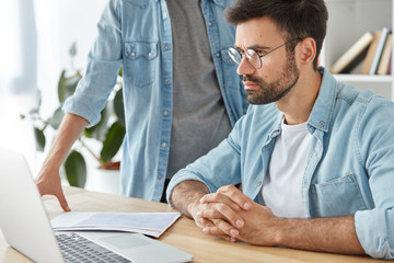 Wall Mural - Indoor shot of two men discuss together work questions, make financial report on laptop computer, work with documents. Male students prepare common project. Cooperation and business concept.