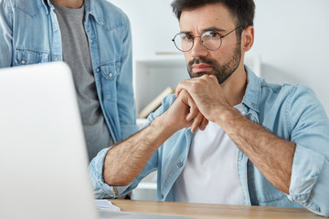 Wall Mural - Handsome attentive unshaven male office worker and his partner stands near, creat business startup project, plan working strategy, use modern laptop computer, collaborate together. Partnership