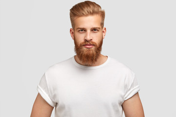 Horizontal shot of prosperous male enterpreneur with appealing look, thick ginger beard, has serious look at camera, listens attentively investors, wears casual t shirt, isolated on white wall