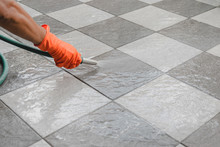 Hand Of Man Wearing Orange Rubber Gloves Is Use A Hose To Clean The Tile Floor.