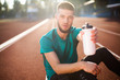 Portrait of young guy dreamily looking in camera showing white sport bottle while spending time on running track of stadium