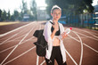 Pretty girl in sport wear dreamily looking in camera with backpack on shoulder and bottle of water in hand on running track of stadium
