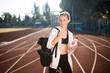 Pretty girl in sporty top and legging dreamily looking aside with backpack on shoulder and bottle of water in hand on racetrack of stadium