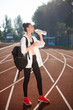 Pretty girl in sporty top and legging with backpack on shoulder drinking water on racetrack of stadium