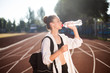 Beautiful girl in sporty wear with backpack on shoulder drinking water on racetrack of stadium