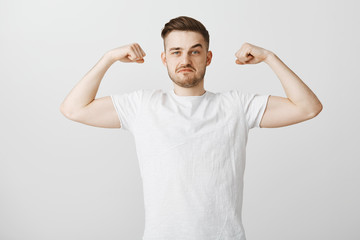 Wall Mural - Strong good-looking male with beard and stylish hairstyle in white casual shirt raising hands showing mascules proudly being in great physical condition working out in gym bregging about biceps