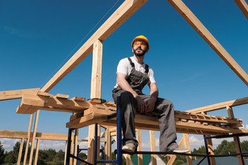 The man builder sits on the edge of the roof of the frame house, in a yellow helmet and gray overalls. The blue sky and clear sunny day.