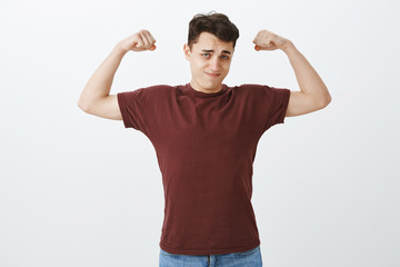 Look how strong I am. Portrait of self-assured handsome european guy in red t-shirt, raising arms and showing muscles with confident expression, bragging about workout and power over gray wall