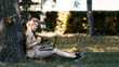 © mark_ka - Young girl student sitting with laptop on grass under tree in park. In one hand holds a cardboard cup of coffee.