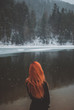 Handsome redhead girl looking in the distance. Snowflakes falling on her hair. Splendid shady black mountain lake. Dreamy winter landscape of Carpathian mountains. Look from behind