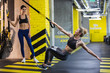 © Yakobchuk Olena - Concentrated slim lady is doing squats while pulling webbing and balancing on heels. Her joyful friend is standing nearby and watching her. Suspension training for woman concept