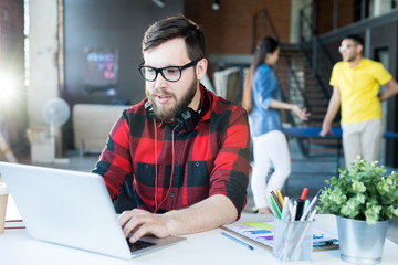 portrait of modern bearded man wearing glasses and red shirt using laptop while working in sunlit of