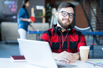 Portrait of smiling bearded man wearing headphones and glasses looking at camera while sitting at desk in modern office of IT developers company, copy space