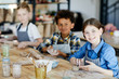 © pressmaster - Happy little girl looking at you while sitting by table and making clay cup with her schoolmates on background
