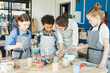 © pressmaster - Group of kids and their teacher leaning over table with gouahes and painting self-made clay items