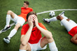 © pressmaster - Stressed or tired footballer sitting on green field and covering his face with hands on background of two mates