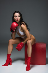 Wall Mural - sexy sportive woman in white swimwear and boxing gloves sitting on red cube on grey