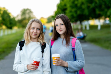 Two teenage girls. Summer in nature. Hands holding cups of coffee and tea. Rest after school behind backpacks. Concept for lunch break, smiling happily.