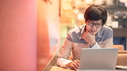 Wall Mural - Young Asian businessman with eyeglasses using laptop computer in cafe. Male entrepreneur searching for inspiration on internet. Business and technology concepts