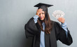 © Krakenimages.com - Young brunette woman over grunge grey wall wearing graduate uniform holding dollars stressed with hand on head, shocked with shame and surprise face, angry and frustrated. Fear and upset for mistake.