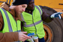 Side View Portrait Of Two Workers, One African-American, Using Digital Tablet Standing Next To Heavy Industrial Truck On Worksite, Copy Space