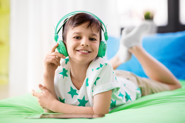 childhood, technology and people concept - smiling boy with tablet pc computer and headphones lying on bed at home