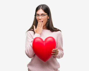 Poster - Young hispanic woman holding heart in love cover mouth with hand shocked with shame for mistake, expression of fear, scared in silence, secret concept