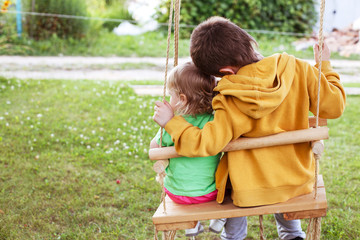 children sitting on a swing in the garden. older brother hugging little sister