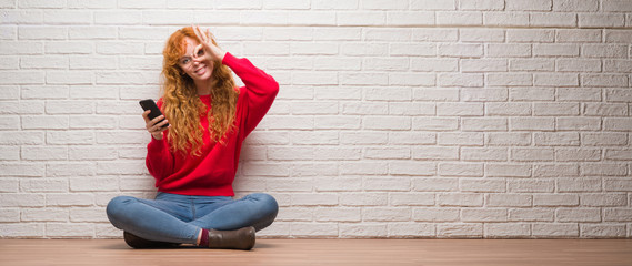 Sticker - Young redhead woman sitting over brick wall talking on the phone with happy face smiling doing ok sign with hand on eye looking through fingers