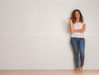 Middle age hispanic woman standing over white brick wall very happy pointing with hand and finger to the side