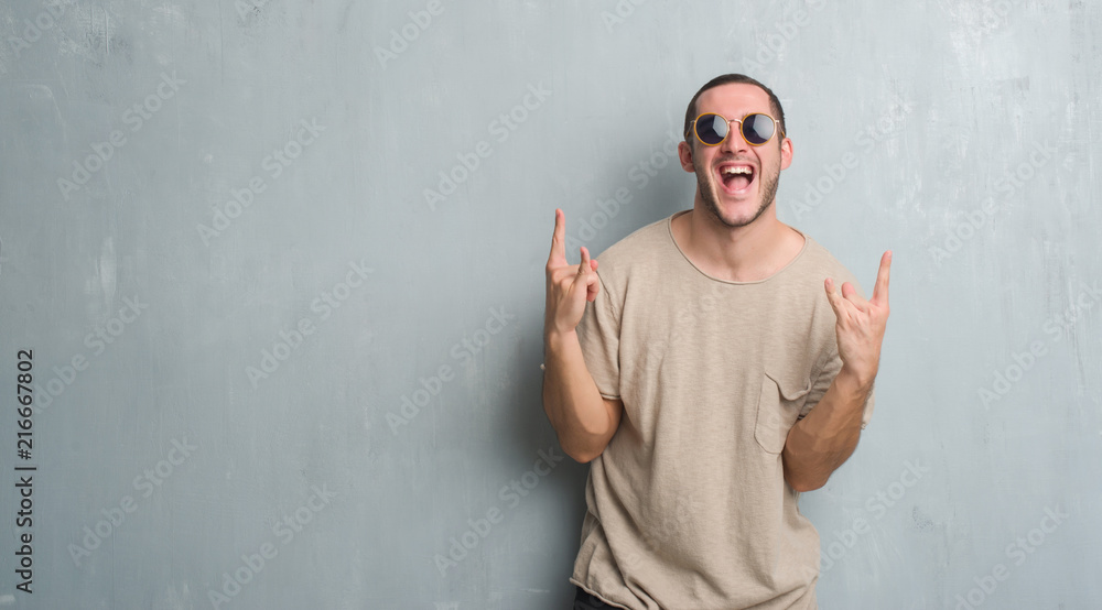 Young caucasian man over grey grunge wall wearing sunglasses shouting with crazy expression doing rock symbol with hands up. Music star. Heavy concept. - obrazy, fototapety, plakaty 