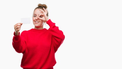 Wall Mural - Young blonde woman holding advertising card with happy face smiling doing ok sign with hand on eye looking through fingers