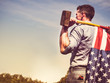 Attractive guy with a sledgehammer and a US flag on his shoulders, looking into the distance against the background of green trees, blue sky and sunset. View from the back. Labour Day concept