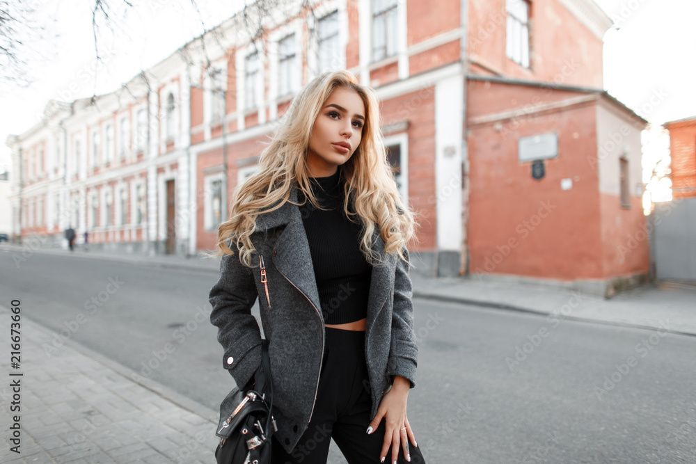 Beautiful young woman model with a bag in a stylish gray jacket walking outdoors in the city - obrazy, fototapety, plakaty 