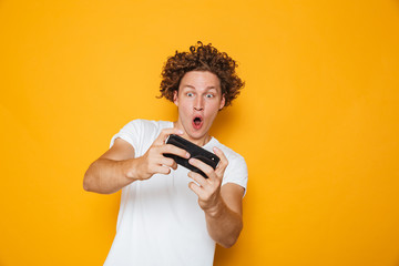 Young brunette guy in casual t-shirt playing online video games using mobile phone, isolated over yellow background