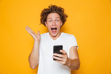 Poster - Happy young man in casual white t-shirt screaming while holding smartphone, isolated over yellow background