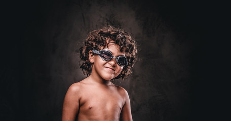 Funny little shirtless boy in swimming goggles in a studio. Isolated on the dark textured background.