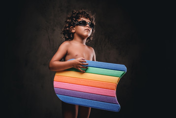 Cute little shirtless boy wears swimming goggles holds a kickboard in a studio. Isolated on a dark textured background.