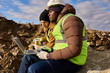 © Seventyfour - Side view  portrait of two miners wearing reflective jackets, one of them African, using laptop in quarry