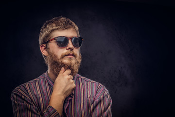 Close-up portrait of a pensive bearded redhead guy wearing sunglasses dressed in an old-fashioned shirt on a dark background.