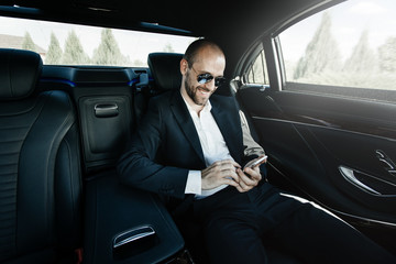 Confident young businessman smiling,using his smartphone while sitting on the back seat of luxury car