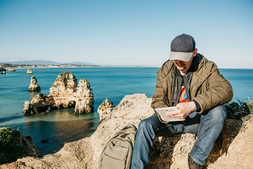 Wall Mural - A young man or a tourist uses a tablet sitting on the Atlantic coast next to the city called Lagos in Portugal. Use of mobile applications or maps or freelancing or remote work or communication.