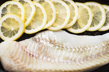 Raw cod fillets with lemon slices on a black baking tray. Top View.