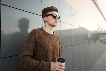 Wall Mural - Fashionable handsome model hipster man with coffee in trendy stylish clothes stands near the wall at sunset